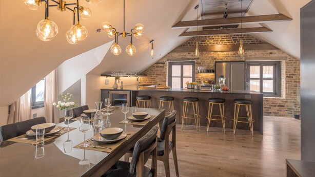 A modern kitchen with exposed brick walls, a large island with stools, and a dining area with a glass table and chairs. - La Maison Kent - Par Aneyro (Québec City)