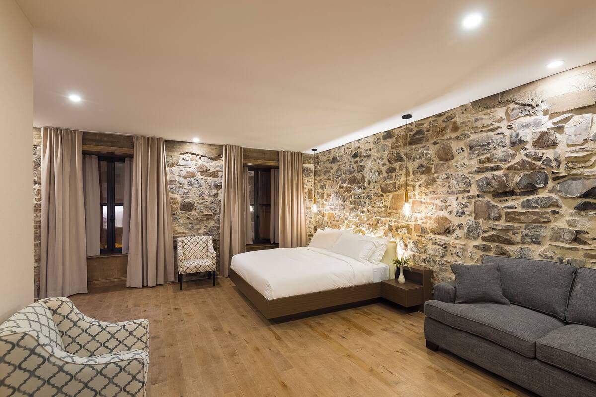Un dormitorio con pared de piedra, cama, sofá gris y sillón estampado.