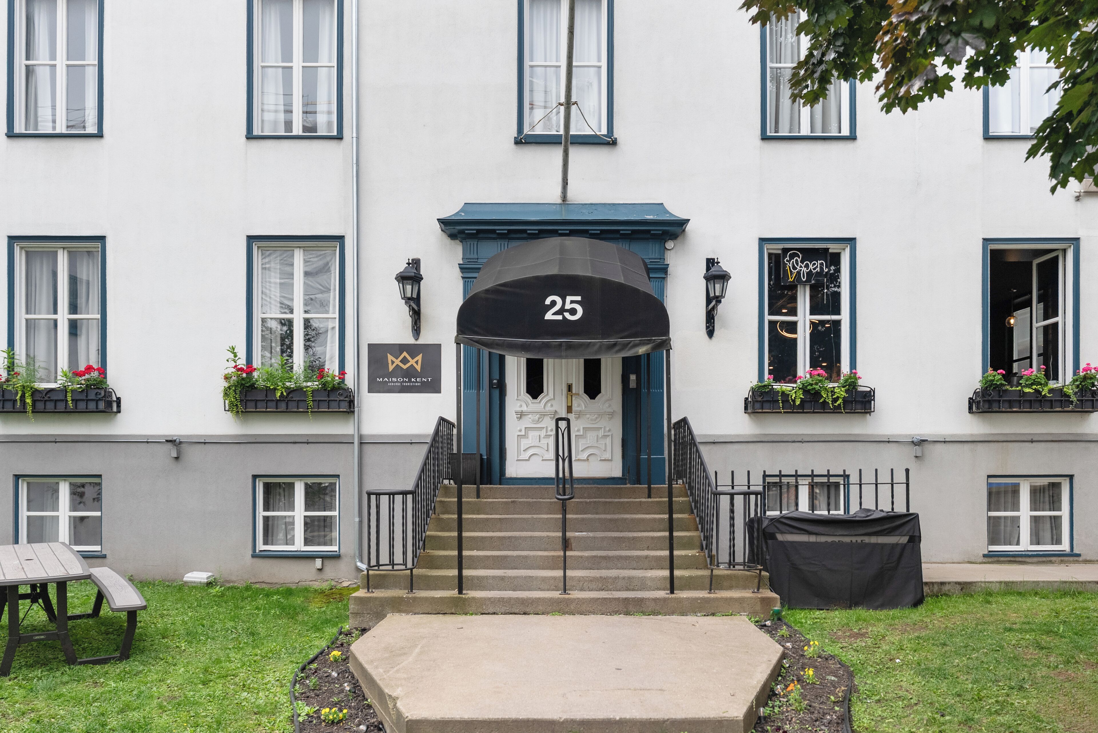 Un immeuble à la façade blanche, portant le numéro 25, avec un auvent bleu au-dessus de l’entrée, des balustrades noires et des jardinières sur les rebords des fenêtres.
