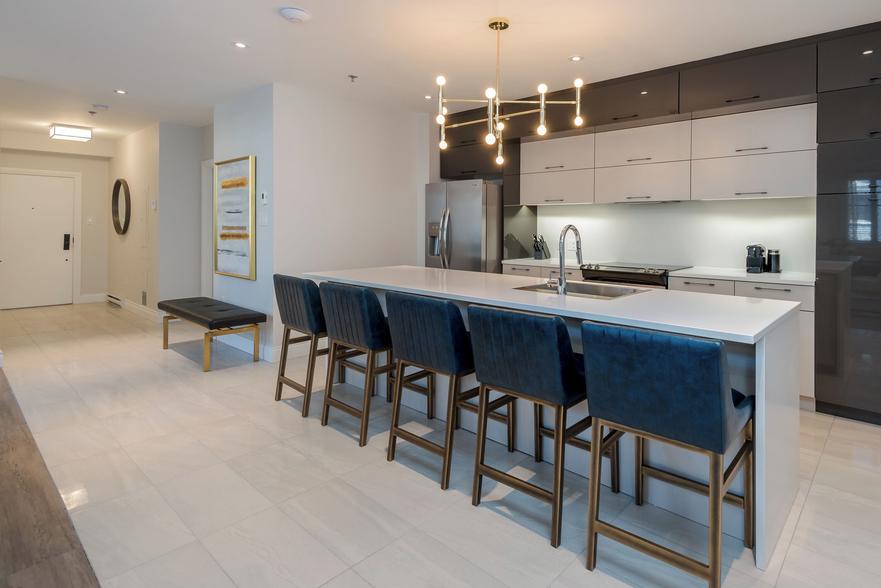 Una cocina moderna con una isla central, taburetes de bar, electrodomésticos de acero inoxidable y una lámpara colgante.