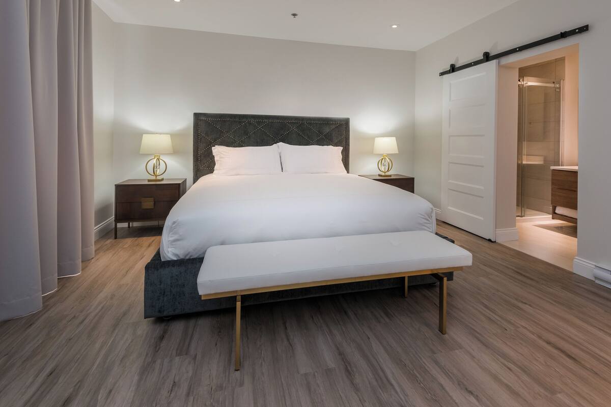 Una habitación de hotel moderna con una cama grande, mesitas de noche con lámparas, una puerta corredera al baño y suelo de madera.