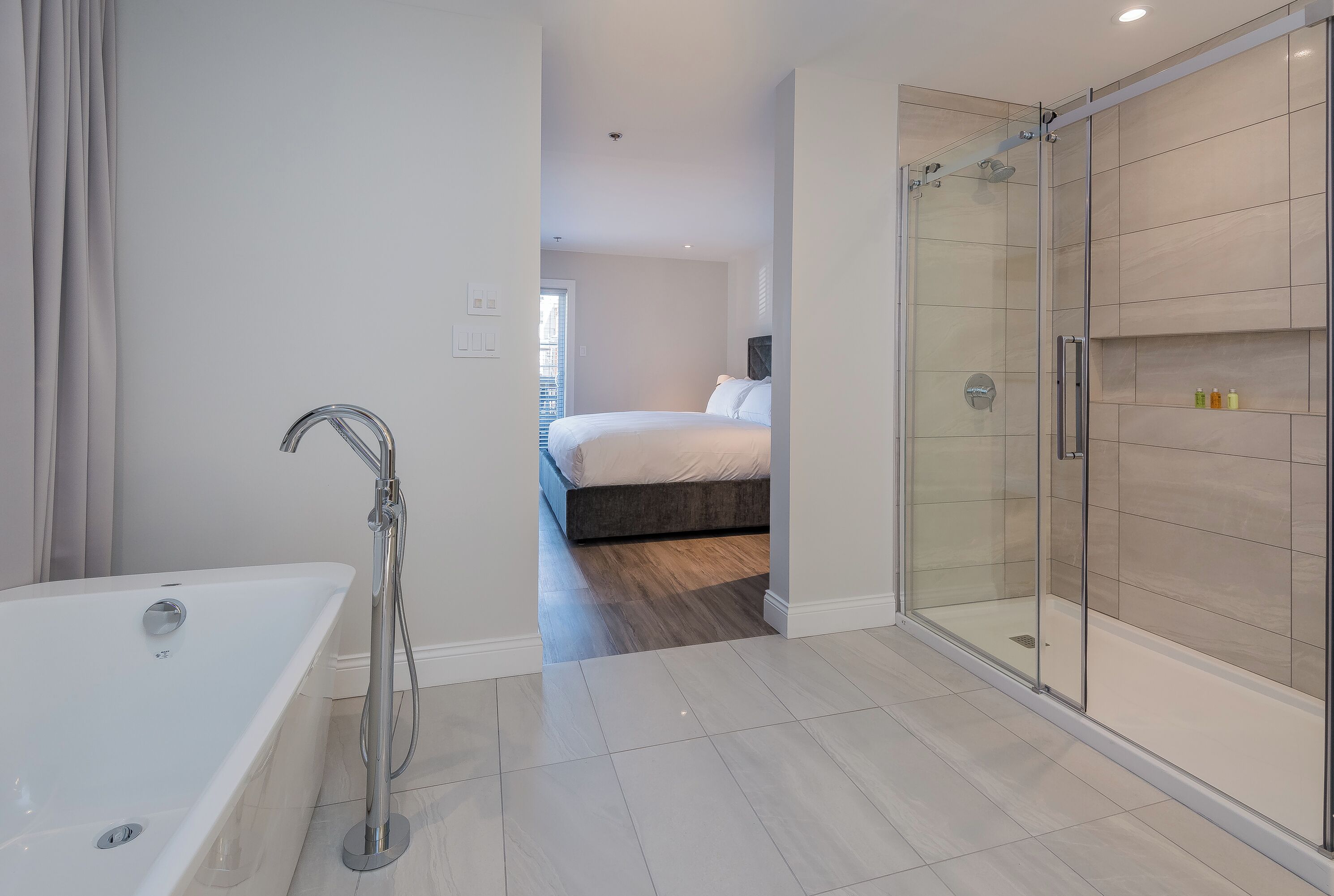 A modern bathroom with a glass-enclosed shower, a standalone bathtub, and a visible bedroom with a bed and a nightstand.