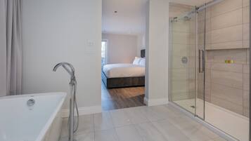 A modern bathroom with a glass-enclosed shower, a standalone bathtub, and a visible bedroom with a bed and a nightstand.