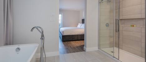 Een moderne badkamer met een glazen douchecabine, een vrijstaand bad en een zichtbare slaapkamer met een bed en nachtkastje.