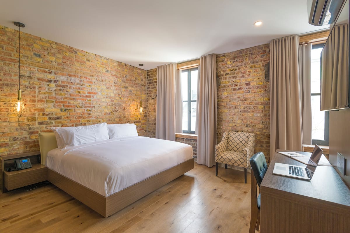 Habitación de hotel con pared de ladrillo, una cama, un escritorio con una computadora portátil, una silla y una ventana con cortinas.