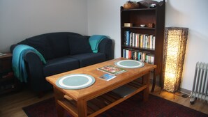 A living room with a dark blue sofa, a wooden coffee table with placemats, a bookshelf filled with books, and a tall floor lamp.