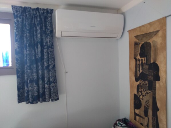A room with a white air conditioner mounted on the wall, a window with blue floral curtains, and a wall-mounted artwork depicting a figure with a bird.