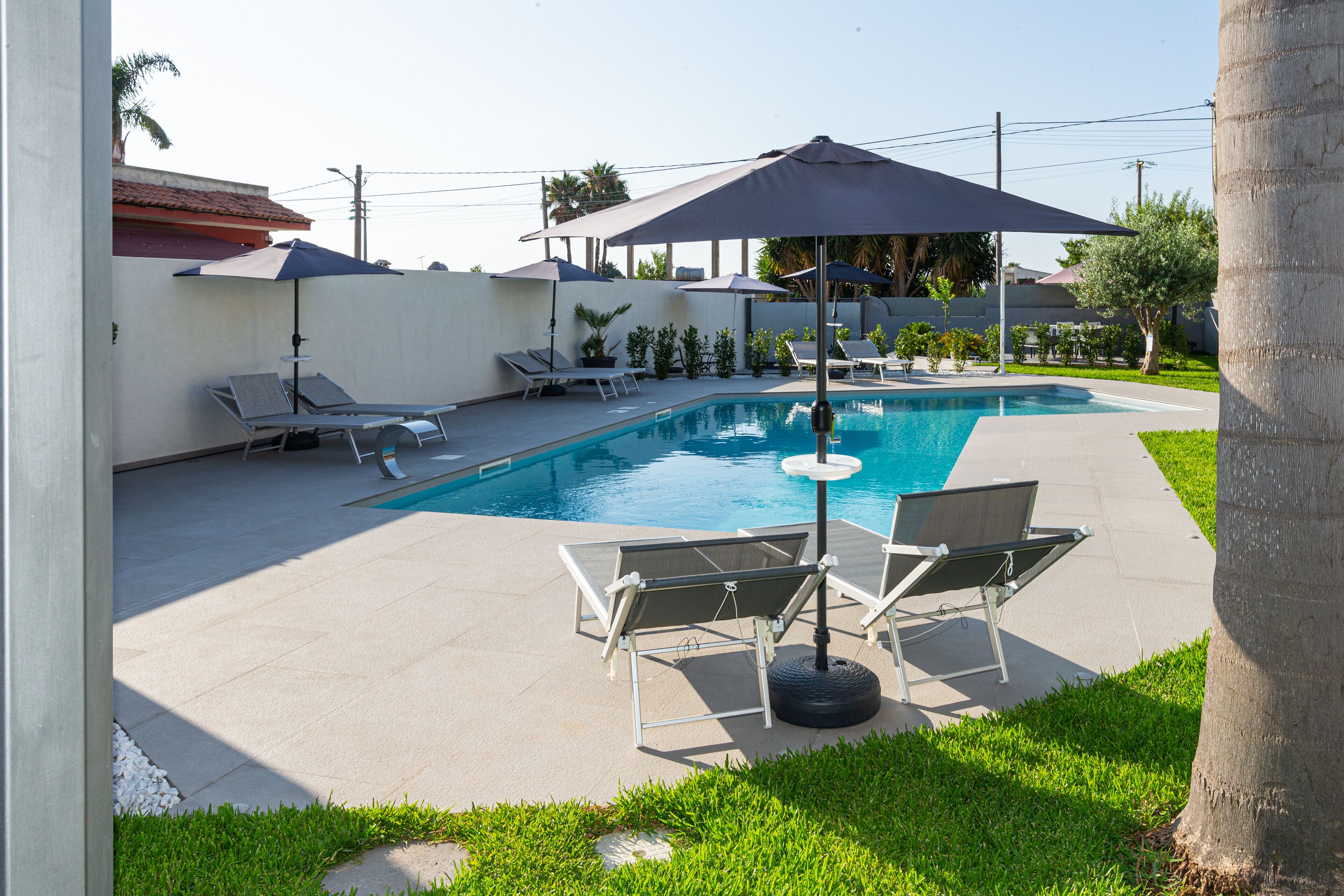 Ein Swimmingpool mit Liegestühlen und Sonnenschirmen, umgeben von einer Betonplattform und viel Grün.