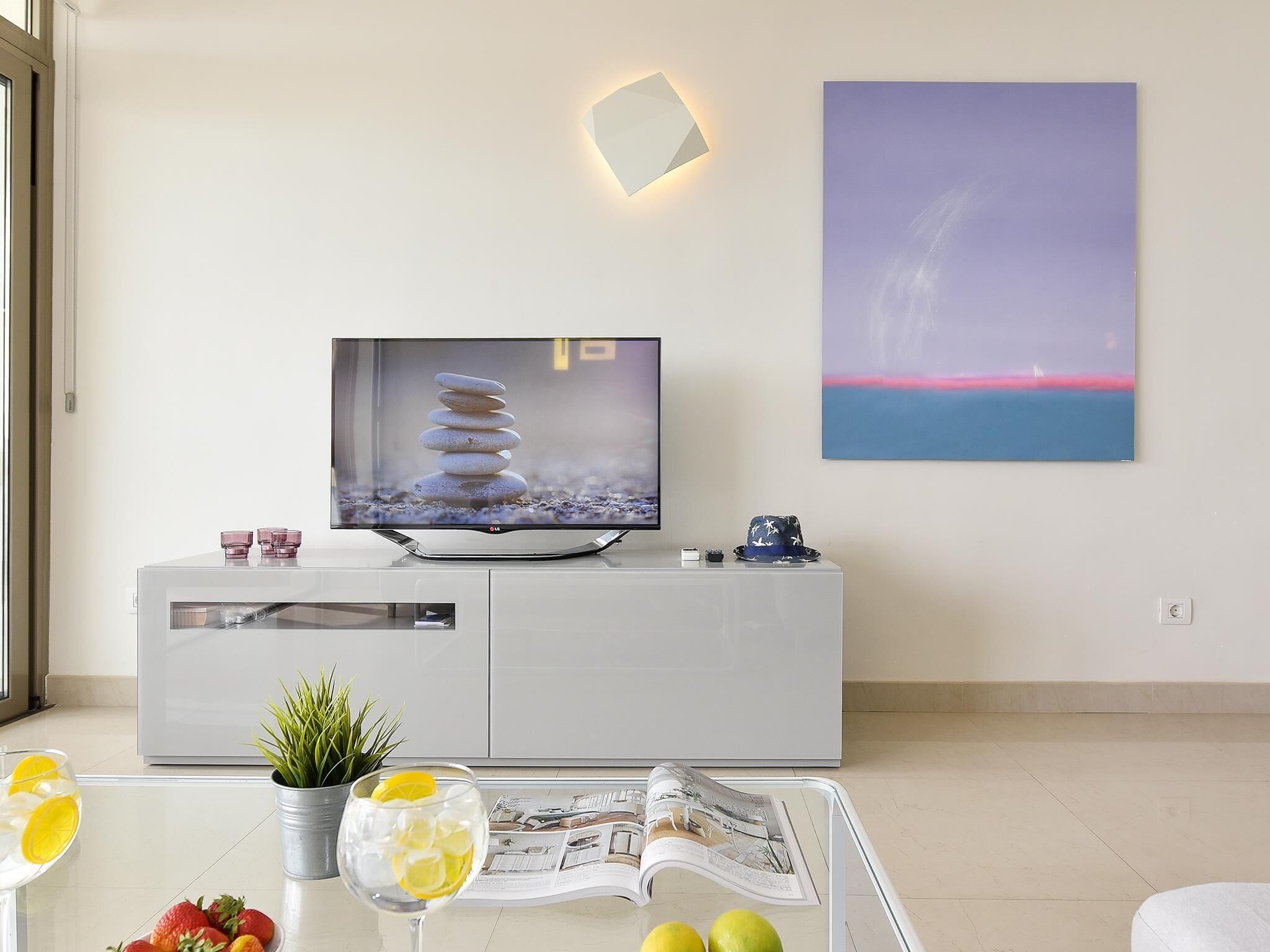 A modern living room with a flat-screen TV, a glass coffee table with magazines, a potted plant, and a decorative wall piece.