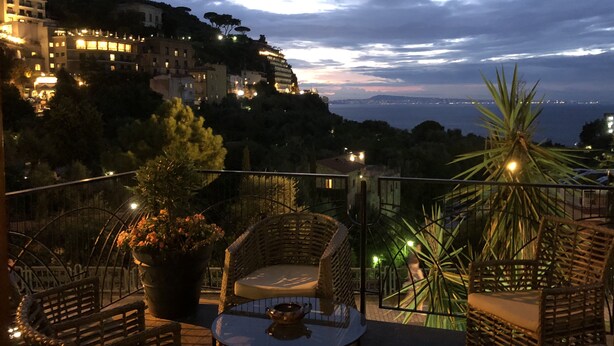 Una terrazza con mobili in vimini, una pianta in vaso e vista su una città costiera al crepuscolo.