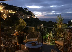 Eine Terrasse mit Korbmöbeln, einem Topfpflanzen und Blick auf eine Küstenstadt im Dämmerlicht.