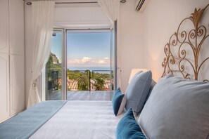 Un dormitorio con cama, almohadas y vistas a un balcón con barandilla y palmeras.