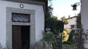 A building with a tiled roof, a wooden door, and a window with decorative bars. There is a paved courtyard with a stone wall and potted plants.