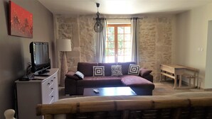 A living room with a stone wall, a television on a cabinet, a sofa with patterned cushions, and a window with curtains.