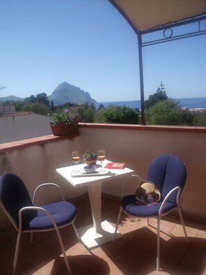 A balcony with two chairs, a table with a bowl, glasses, and a book. A mountain and the sea are visible in the background. - Red bougainvillea - sea view - 2 steps from the bay.  Parking - WiFi     (San Vito Lo Capo)
