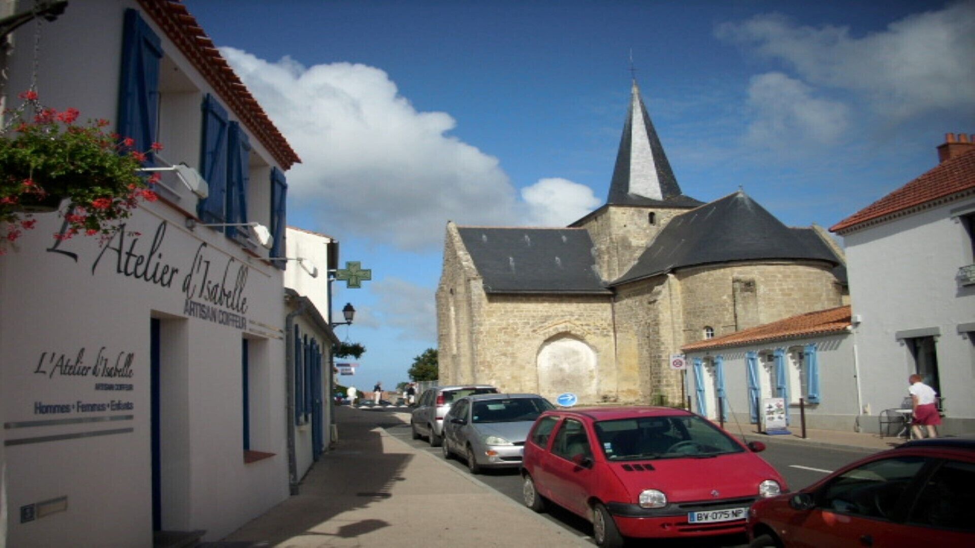 A narrow street with parked cars, a church with a tall steeple, and a building with a sign reading "Atelier d'Isabelle".