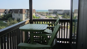 Un balcon avec une table et des chaises, donnant sur un parking et sur des immeubles, avec vue sur l’océan.