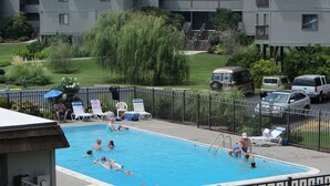 Une piscine entourée d’une clôture métallique, où l’on voit des gens nager et se détendre au bord de l’eau.