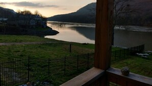 A riverside view with a wooden deck, a fenced grassy area, and a house in the background.