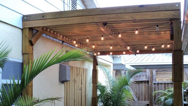 Une pergola en bois, décorée de guirlandes lumineuses, entourée de plantes en pot et d’une clôture en bois.