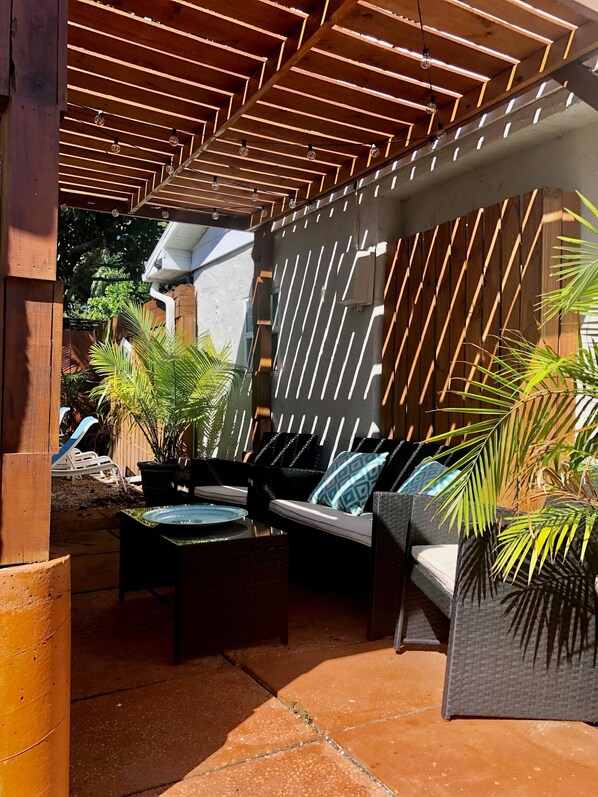 An outdoor patio area with a wooden pergola, cushioned seating, and potted plants.