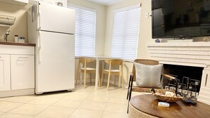 Une cuisine avec un réfrigérateur blanc, un coin repas avec deux chaises, un téléviseur fixé au-dessus d’une cheminée, et une table basse ronde sur laquelle se trouvent un panier et une télécommande.