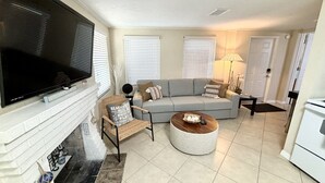 Un salon équipé d’un téléviseur à écran plat, d’un canapé gris, d’un fauteuil en osier, d’une table basse et d’une cheminée.