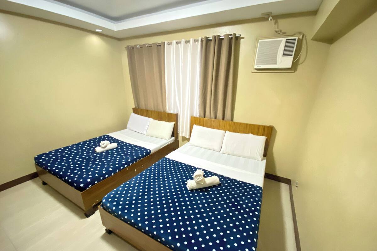 Quarto de hotel com duas camas, cada uma com colcha azul de bolinhas e almofadas brancas. Há um ventilador de teto e uma janela com cortinas bege.