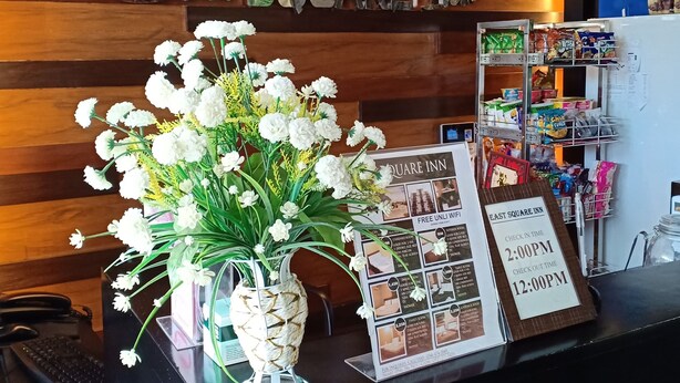 A reception desk with a wooden backdrop, a vase of white flowers, and a signboard with check-in and check-out times. - East Square Inn (Bacolod)