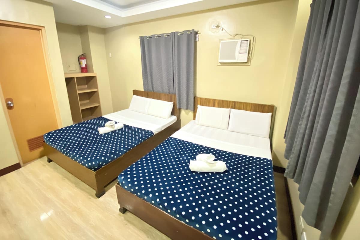Quarto de hotel com duas camas, cada uma com colcha de bolinhas azuis e almofadas brancas, cabeceira de madeira, uma pequena prateleira com um objeto vermelho, cortina cinza e ar condicionado fixado na parede.