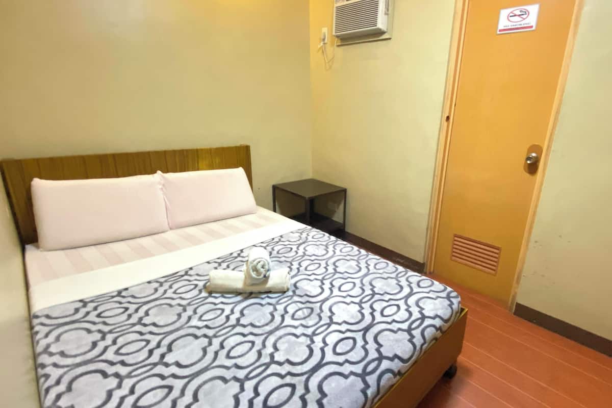 Quarto de hotel com cama, mesa de cabeceira, ar condicionado na parede e porta com placa de proibido fumar.