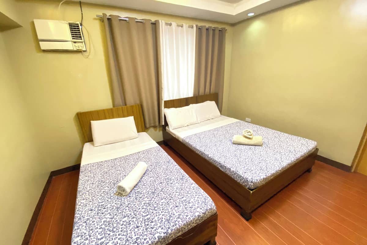 Quarto de hotel com duas camas, cada uma com roupa de cama e almofadas estampadas, cabeceira de madeira, ventilação no teto e uma janela com cortinas.