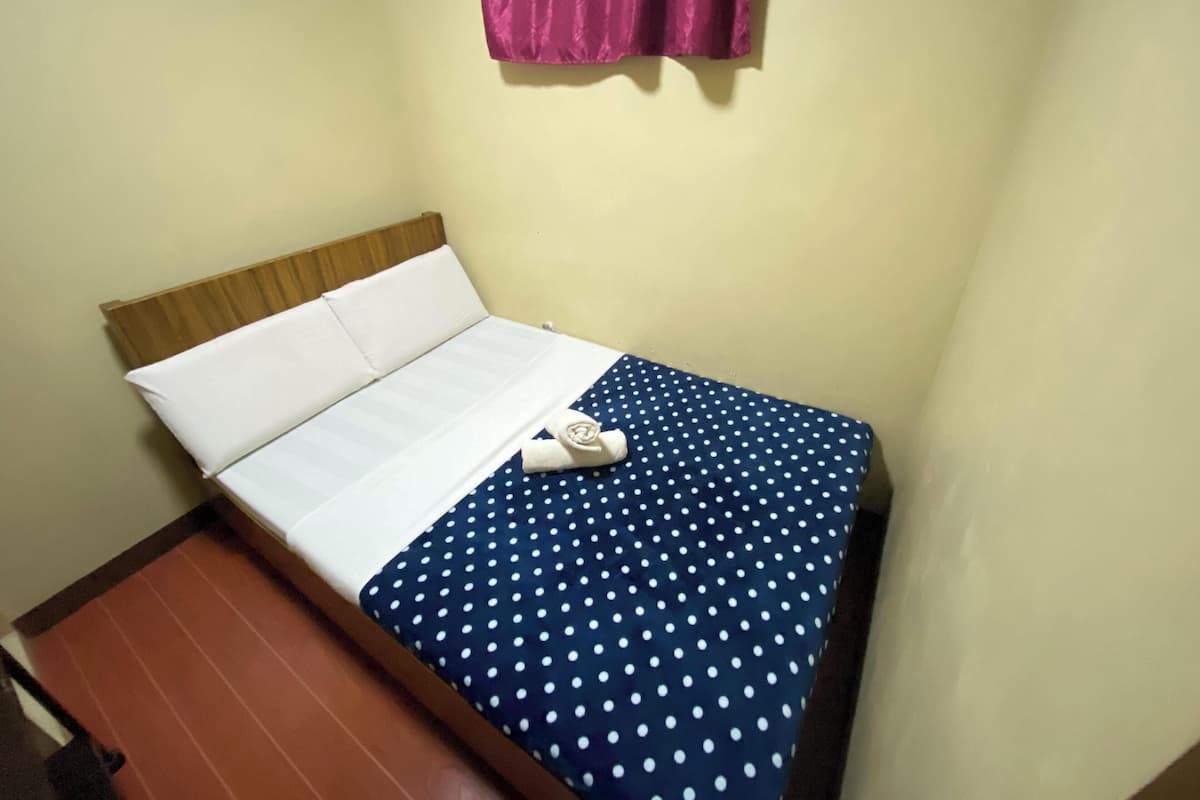 Uma cama de solteiro com roupa de cama branca e uma colcha azul com bolinhas, uma cabeceira de madeira e uma cortina rosa.