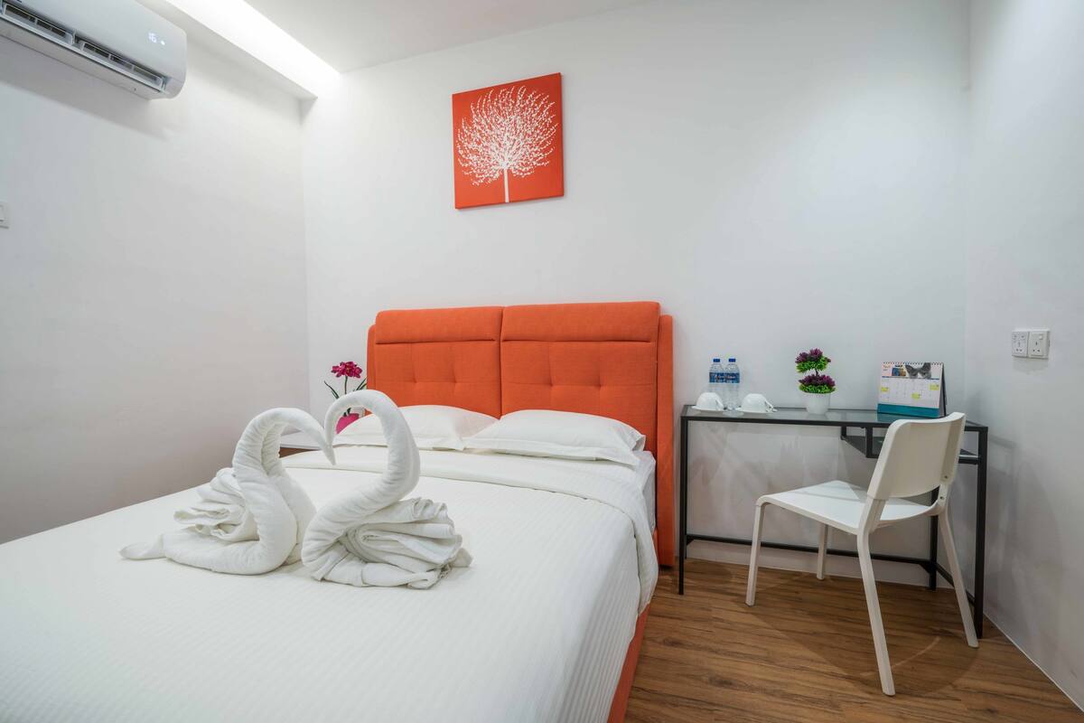 A hotel room with a bed, a desk, a chair, a wall-mounted air conditioner, and a red artwork.