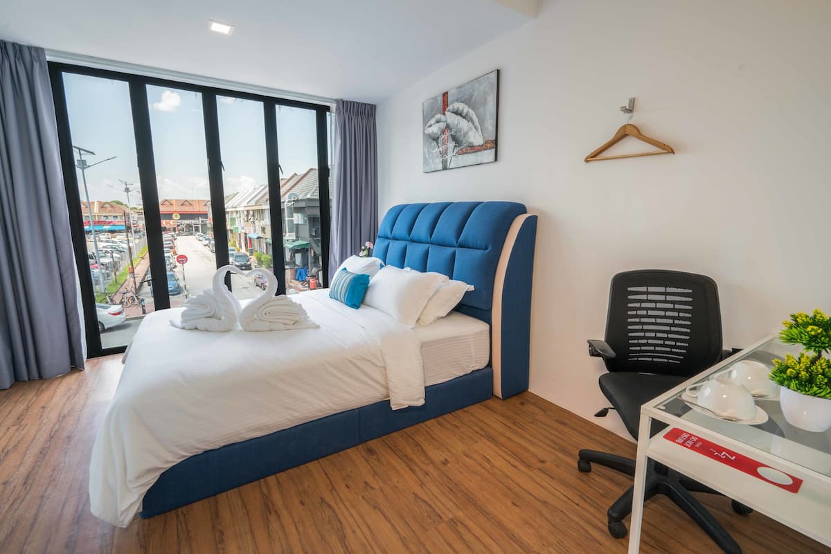 A hotel room with a large bed, a desk with a chair, a hanging coat rack, and a view of the street through a large window.