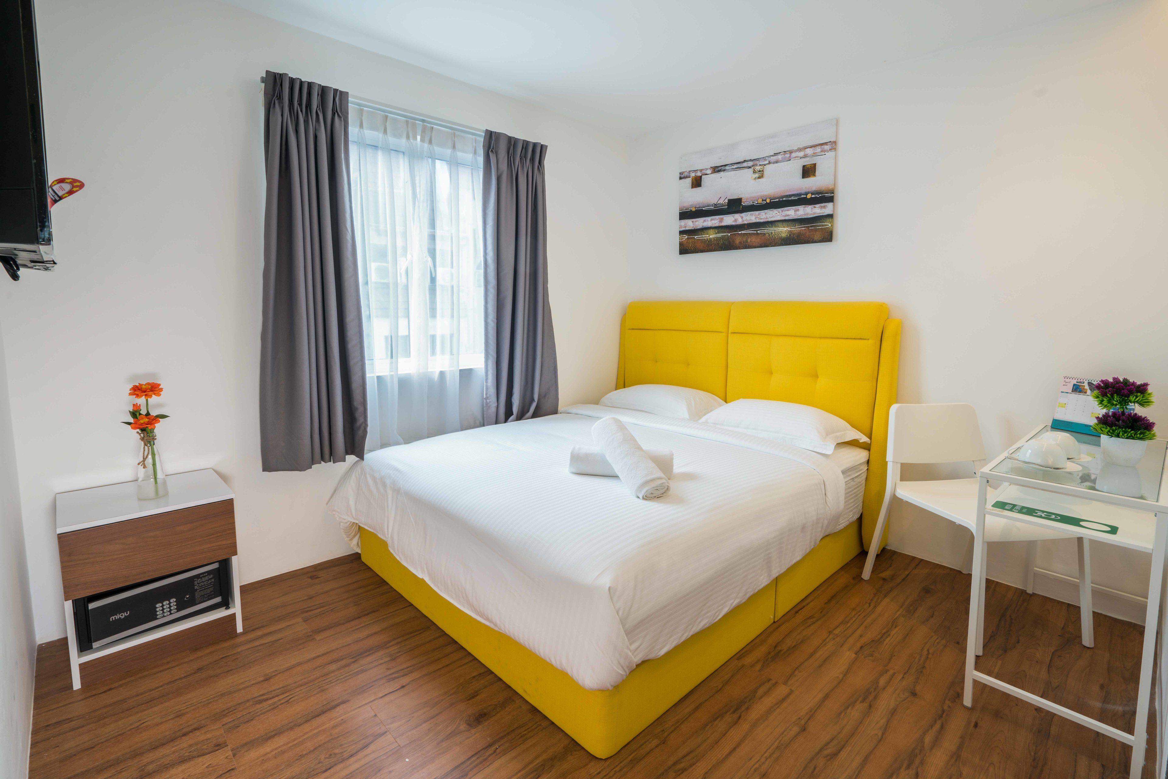 Quarto de hotel com cama amarela, roupa de cama branca, mesa de cabeceira com abajur, pequena mesa com um vaso e janela com cortinas.