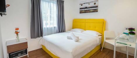 Quarto de hotel com cama amarela, roupa de cama branca, mesa de cabeceira com abajur, pequena mesa com um vaso e janela com cortinas.