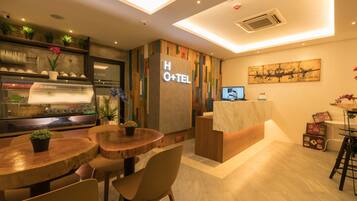 A hotel reception area with a wooden reception desk, a glass display case, and a seating area with round tables and chairs.