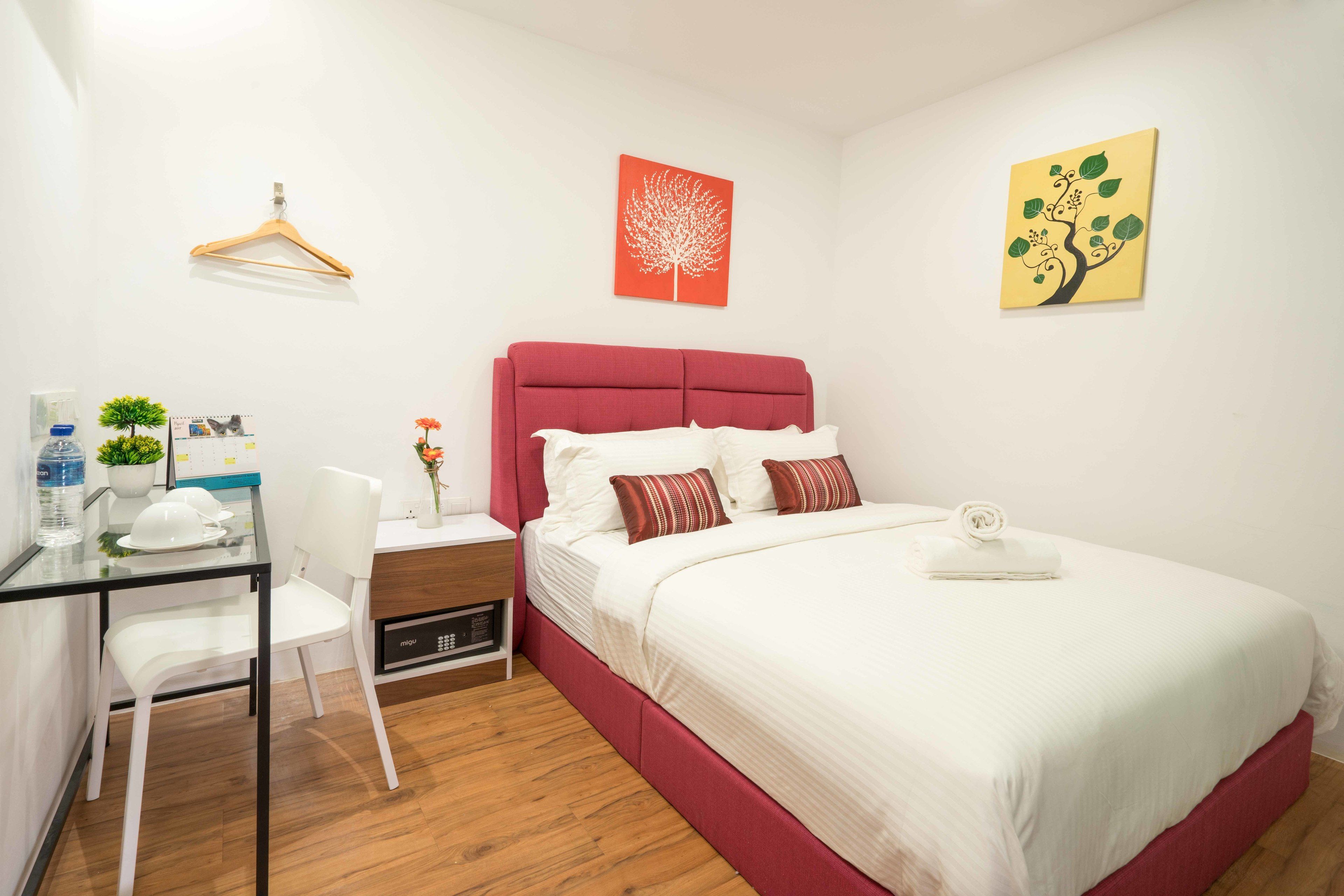 Habitación de hotel con cama, escritorio con silla, una mesita con planta y un perchero en la pared.