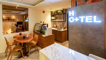 Uma área de recepção de hotel com uma mesa de mármore, cadeiras de madeira, um balcão de recepção e uma placa com a inscrição "HOTEL+TEL".