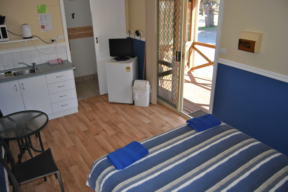 A compact hotel room with a kitchenette, a bed with striped bedding, a small table with chairs, and a view through a glass door.