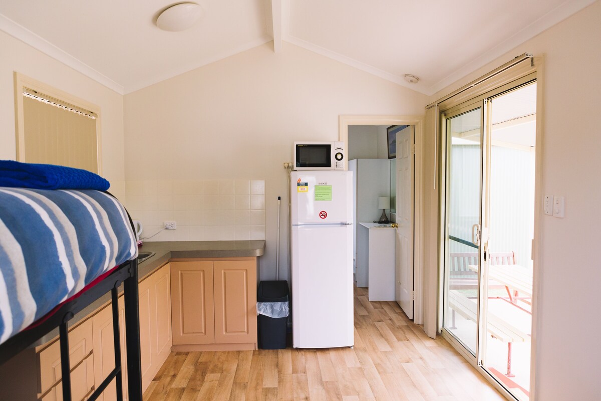 A compact kitchen with a microwave, refrigerator, and a small trash bin. There is a bed with a striped blanket and a wooden floor.