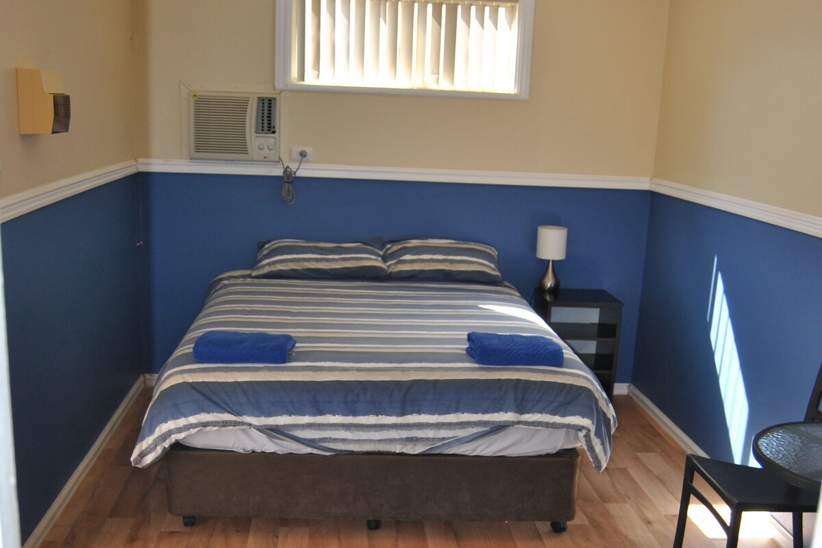 A bedroom with a double bed, a bedside table with a lamp, a chair, a window with blinds, and an air conditioning unit.