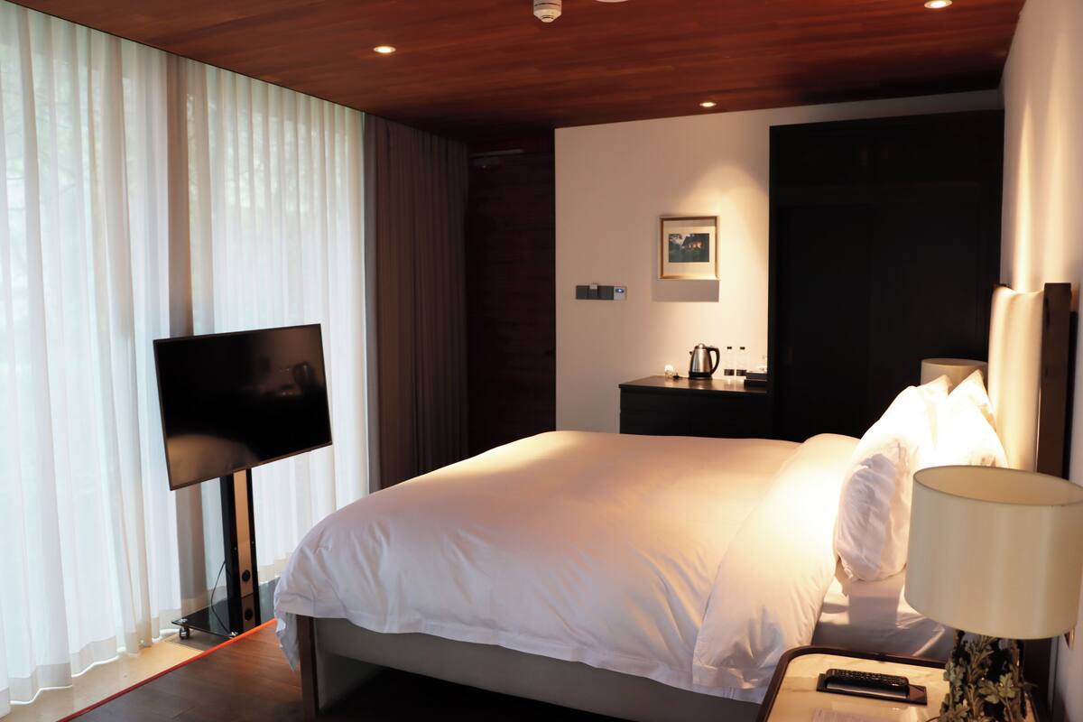 Una habitación de hotel moderna con una cama grande, un televisor de pantalla plana, una mesita de noche con lámpara y una mesita auxiliar.