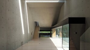 Ein großzügiger Innenraum mit hoher Decke, Betonwänden und Holzboden. Eine Treppe führt zu einer erhöhten Ebene.
