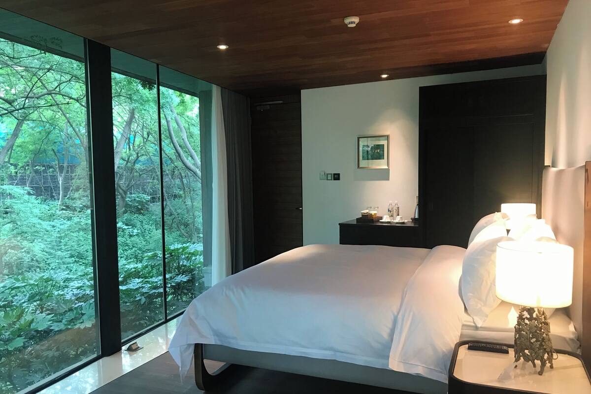 Un dormitorio moderno con una cama grande, una mesita de noche con lámpara y vistas a la vegetación a través de amplias ventanas.