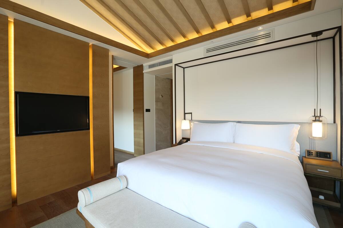 Una habitación de hotel moderna con una cama grande, un televisor de pantalla plana montado en la pared y una mesita de noche con una lámpara.
