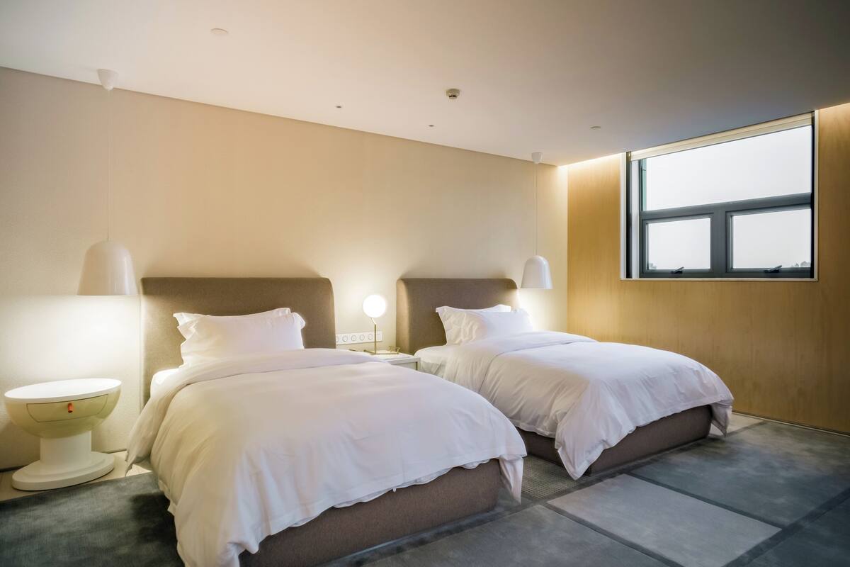 Habitación de hotel con dos camas, lámparas de noche, un espejo redondo y un ventanal grande.