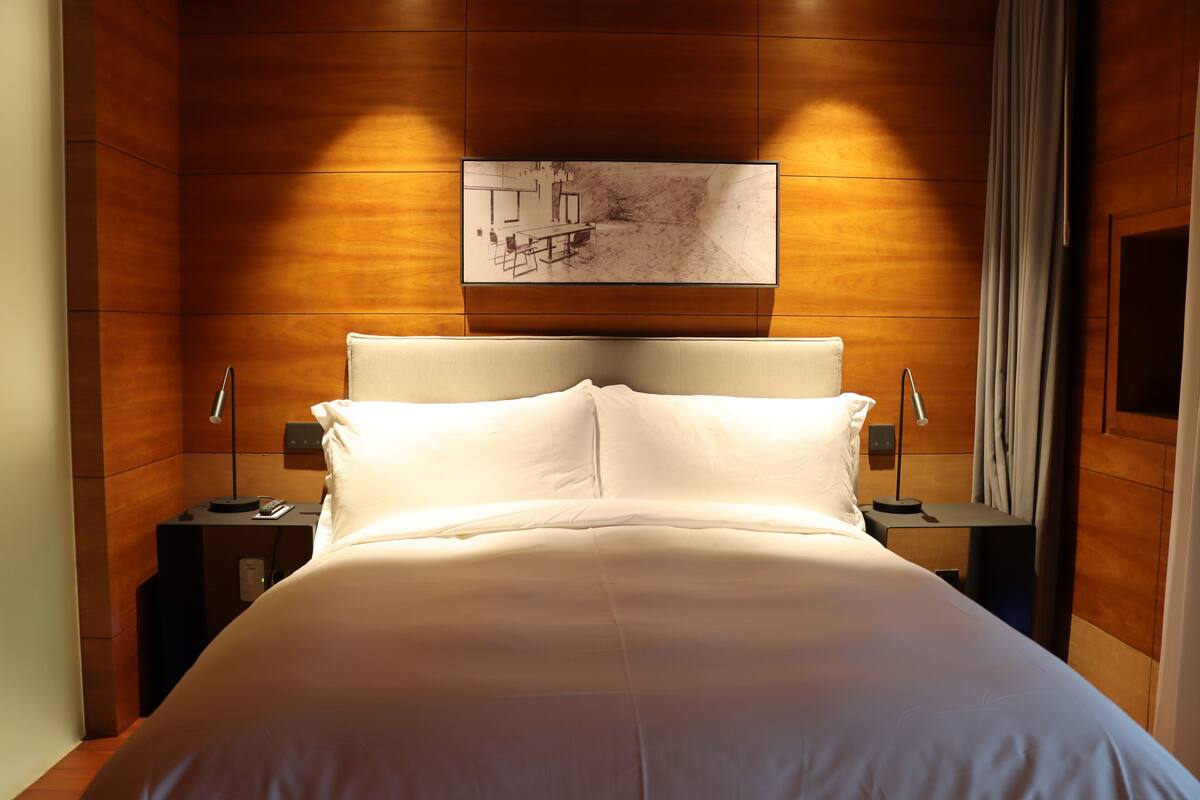 Una cama bien hecha con ropa blanca, cabecera de madera y una obra de arte colgada en la pared.