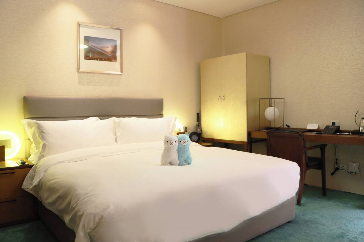 Habitación de hotel con una cama grande, un escritorio y una silla. Se observa un cuadro en la pared y una lámpara sobre el escritorio.